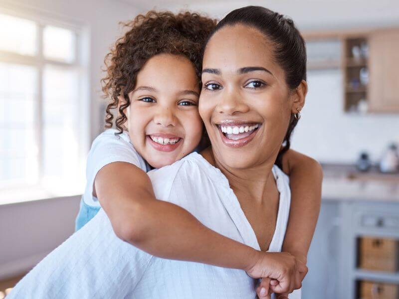 A woman and a child are smiling as the child hugs the woman from behind